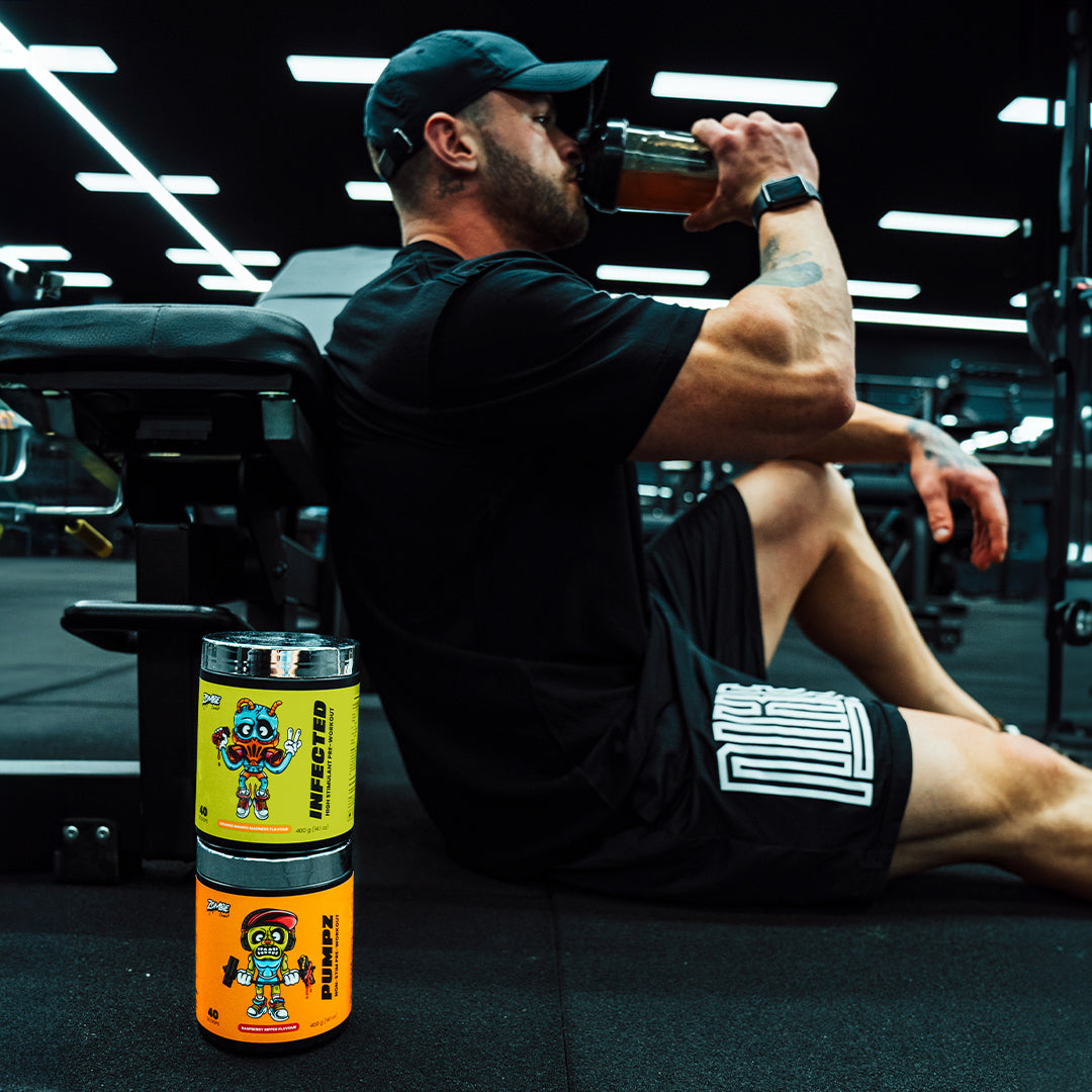 Man dressed in black gymwear sitting next to a bench in a gym drinking out of a black shaker with Zombie Labs Infected Preworkout stacked on top of Zombie Labs Pumpz Preworkout