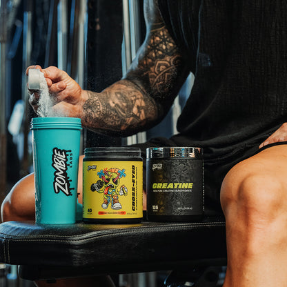 Man sitting on gym bench tipping a scoop of Zombie Labs Cross-Eyed Preworkout into a blue Zombie Labs shaker, along with a tub of Zombie Labs Creatine, ready to be mixed