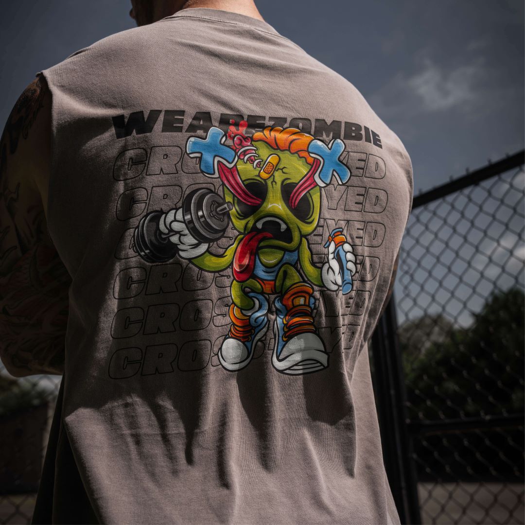 A man with tattooed arms standing outdoors, showing the back of his grey Zombie Labs sleeveless tank top featuring a colorful cartoon character and 'WEAREZOMBIE CROSSEYED' text.