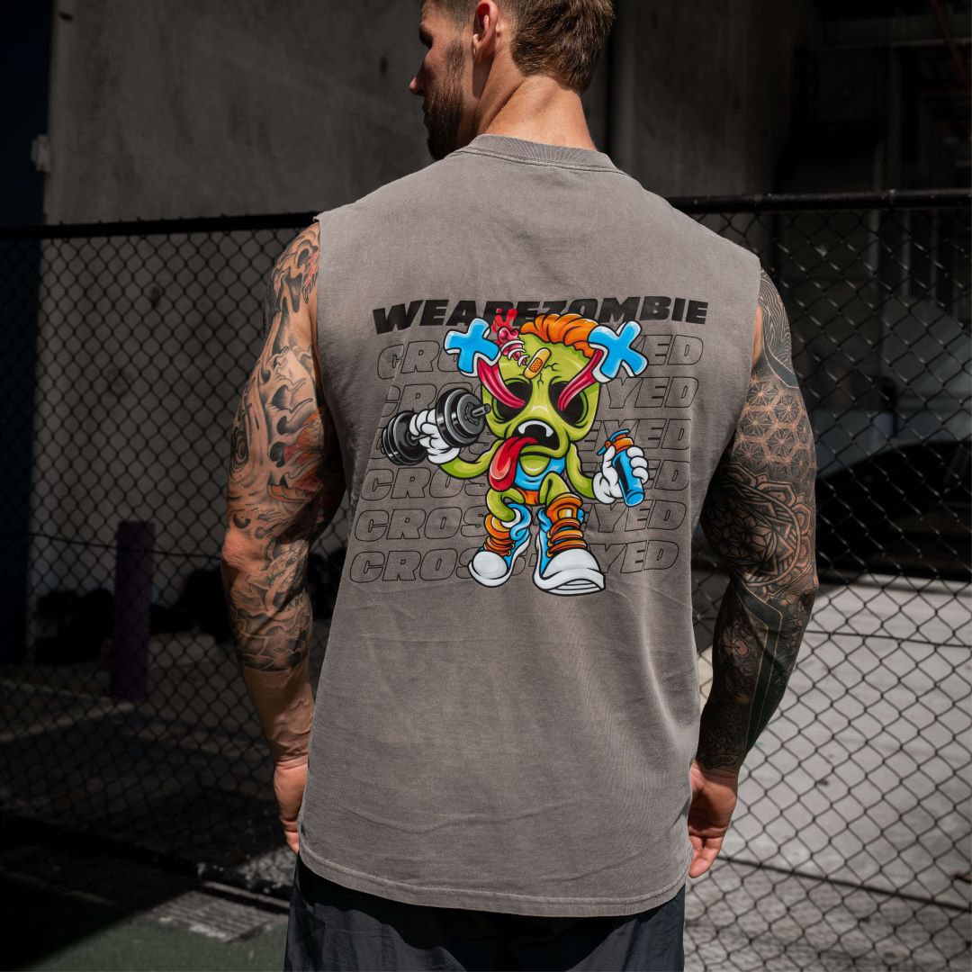 A muscular man with tattooed arms lifting weights in a gym, wearing a grey Zombie Labs sleeveless tank top with a colorful cartoon character graphic and 'WEAREZOMBIE CROSSEYED' text on the back.