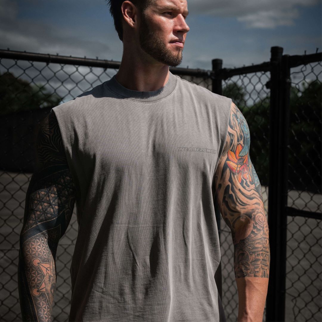 A muscular man with tattooed arms wearing a grey sleeveless tank top from Zombie Labs, standing outdoors next to a chain-link fence under a cloudy sky