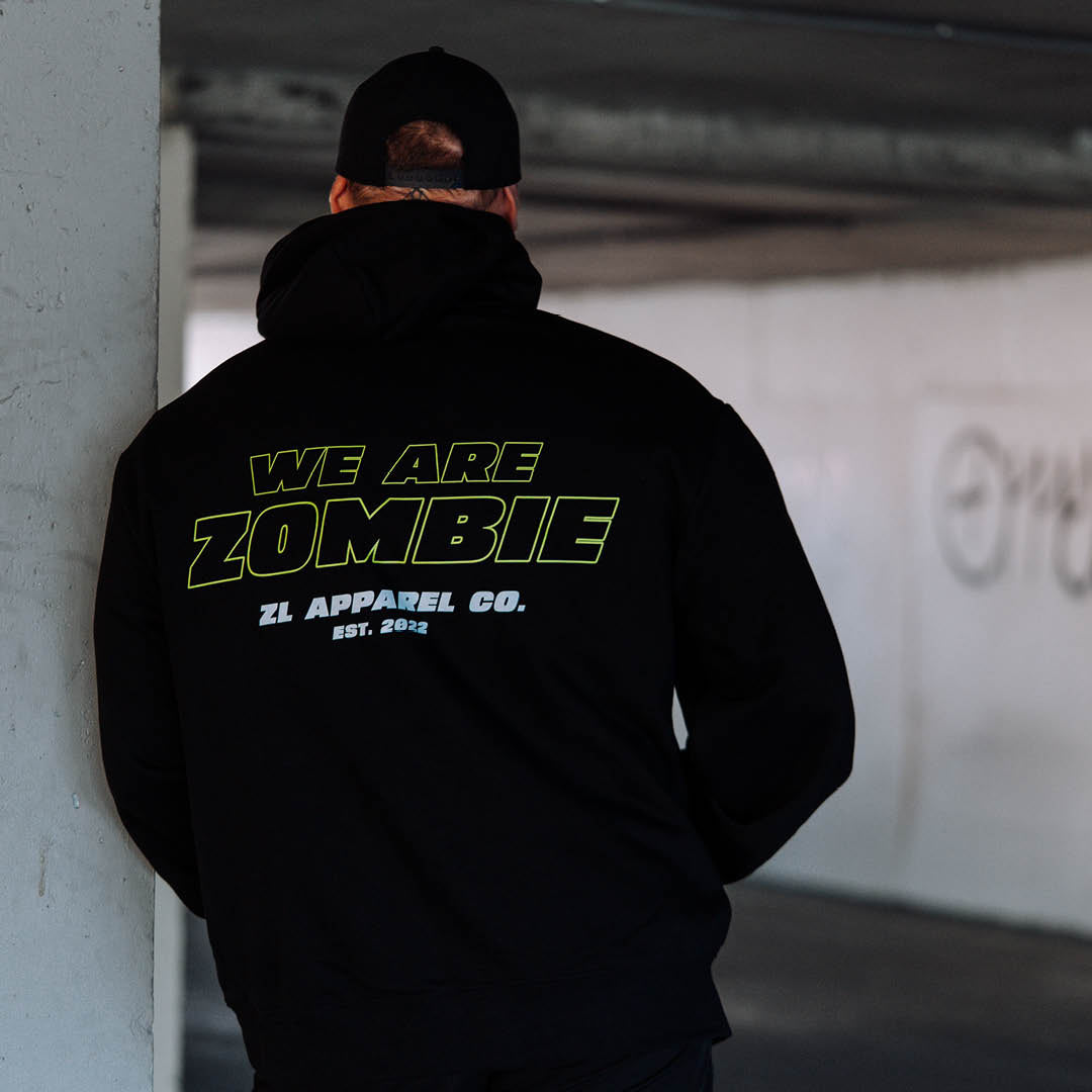 A man leaning against a concrete structure wearing a black Zombie Labs hoodie with WE ARE ZOMBIE on the back. 